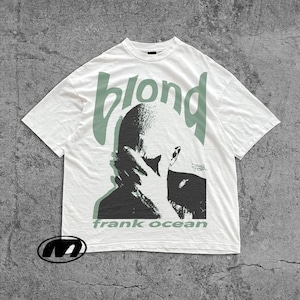 Puede incluir: Camiseta blanca con un gráfico en blanco y negro de una persona, la palabra "blond" en verde encima de la imagen y "frank ocean" en verde debajo. La camiseta tiene mangas cortas y cuello redondo.