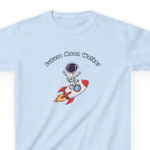 Op de afbeelding: Lichtblauw T-shirt met een cartoon astronaut op een raket. De raket heeft rode en gele vlammen. De tekst "Future Moon Walker" is boven het astronaut en raket ontwerp gebogen.