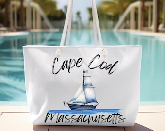 以下が含まれることがあります： 白いトートバッグに、青い帆船と「Cape Cod Massachusetts」の文字がプリントされています。バッグはタイル張りの床に置かれ、プールに隣接しています。