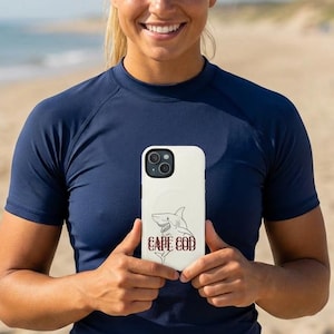 May include: A white iPhone case featuring a shark illustration and the words "CAPE COD" in a dark red font. The case is held in front of a person wearing a navy blue shirt, with a beach and ocean in the background.