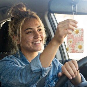 May include: A rectangular car air freshener with a daisy floral design. The air freshener features white daisies with yellow centres against a pink watercolour background. The air freshener is hanging from the rearview mirror.