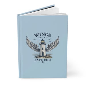 Op de afbeelding: Een lichtblauw notitieboek met de tekst "WINGS NECK CAPE COD" en een illustratie van een vuurtoren met vleugels. Het notitieboek is open en toont witte gelinieerde pagina's.