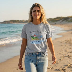 Może przedstawiać: Szara koszulka z grafiką latarni morskiej Nauset Light w Cape Cod, MA. Latarnia jest czerwono-biała, umieszczona na zielonym wzgórzu z niebieską wodą i niebem. Koszulka jest połączona z niebieskimi dżinsami.