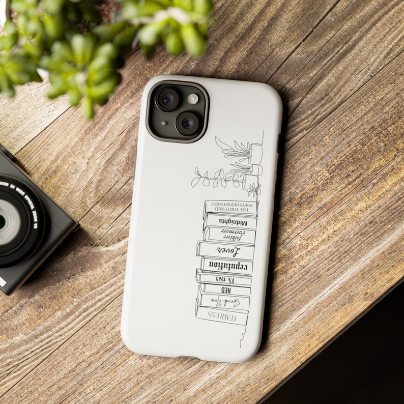 May include: White phone case with a black line drawing of a stack of books with titles like "Fearless", "Red", "12. 1995", "Revolution", "Lover", "Forever", "Covenant", "Midnights", and "The Tormented", with a small plant on top.