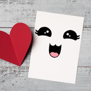 May include: A white card with a black and white cartoon face with big eyes and a wide smile. The card is next to a red paper heart.