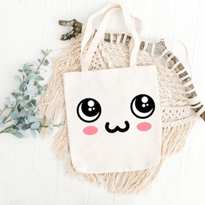 May include: A white canvas tote bag with a cute cartoon face printed on the front. The face has large eyes, a small mouth, and pink cheeks.