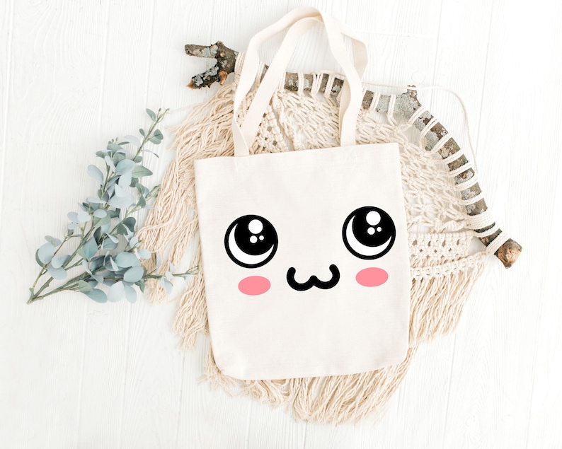 May include: A white canvas tote bag with a cute cartoon face printed on the front. The face has large eyes, a small mouth, and pink cheeks.