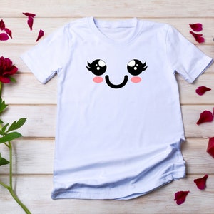 May include: White t-shirt with a cartoon face printed on the front. The face has large black eyes, eyelashes, pink cheeks, and a black smile.
