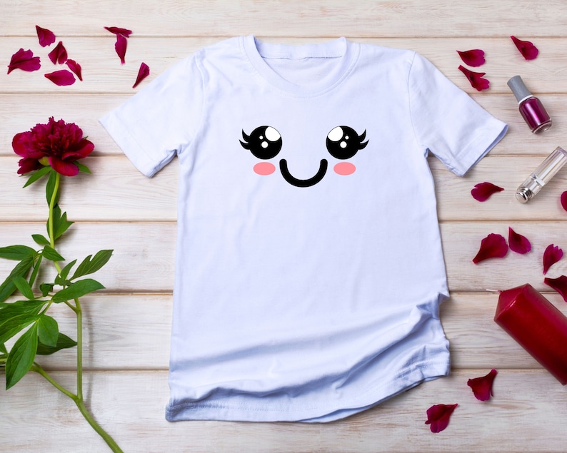 May include: White t-shirt with a cartoon face printed on the front. The face has large black eyes, eyelashes, pink cheeks, and a black smile.