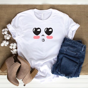 May include: White t-shirt with a cartoon face design featuring black eyes with hearts and pink cheeks. The face is blowing a kiss.