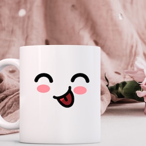 May include: White ceramic mug with a cartoon face with closed eyes and a tongue sticking out. The face has pink cheeks. The mug is on a white surface with a pink fabric and a pink flower in the background.