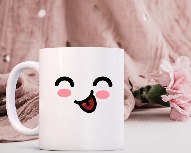 May include: White ceramic mug with a cartoon face with closed eyes and a tongue sticking out. The face has pink cheeks. The mug is on a white surface with a pink fabric and a pink flower in the background.