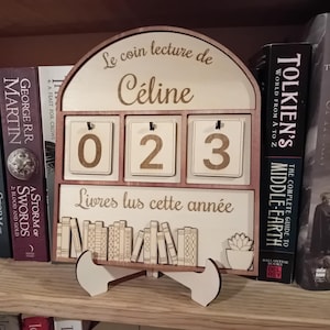 Peut inclure: Calendrier en bois pour coin lecture avec le nom "Céline" et le texte "Livres lus cette année". Le calendrier affiche le nombre "023". Le calendrier est sur une étagère en bois avec des livres.