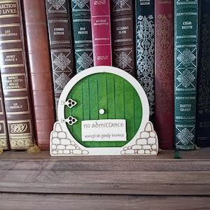 Peut inclure: Une porte verte ronde en bois avec l'inscription "no admittance except on party business" sur une pancarte. La porte est flanquée de structures en forme de pierre et placée devant des livres anciens.