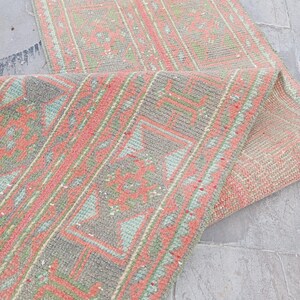 Puede incluir: Una alfombra rectangular tipo pasillero con un patrón tejido en tonos rosa coral, verde salvia y gris apagado. El diseño incluye bordes geométricos y motivos centrales. La alfombra está doblada para mostrar la textura del reverso.