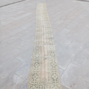 May include: A long, rectangular rug with a faded, vintage appearance. The rug features a central beige stripe flanked by patterned borders in shades of green, beige, and gray. The rug's design includes geometric and floral motifs.