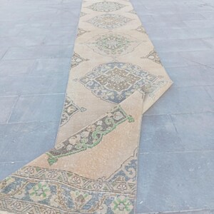 May include: A long, rectangular beige runner rug with an intricate geometric pattern in shades of blue, green, and brown. The rug is partially rolled up, revealing the detailed design. The rug is likely made of wool or a similar material.