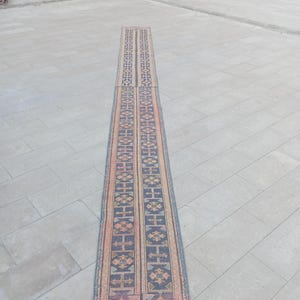 May include: A long, narrow rug with a geometric pattern in shades of blue, orange, and pink. The rug has a central design with repeating shapes and borders. The rug is placed on a stone surface.