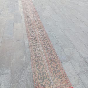 Könnte beinhalten: Ein langer, rechteckiger Teppich mit einem sich wiederholenden geometrischen Muster in Rot-, Braun- und Beigetönen. Der Teppich hat einen roten Fransen an den Enden und wird auf einer grauen Fliesenoberfläche präsentiert. Das Design des Teppichs weist ein zentrales Feld mit einem symmetrischen Muster auf.