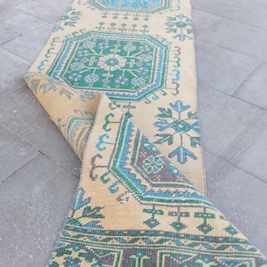 May include: A long, narrow runner rug featuring a beige base with repeating geometric patterns in shades of green, blue, and dark grey. The design includes octagonal motifs and floral-like accents. One end of the rug is folded over itself.
