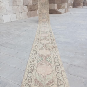 May include: A long, narrow runner rug featuring a faded, muted color palette of beige, soft pink, and sage green. The rug displays a repeating geometric and floral pattern with a decorative border, extending vertically across the floor surface.