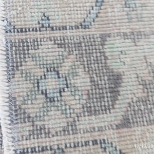 May include: A woven rug featuring a detailed pattern in muted shades of beige, sage green, and charcoal gray. The edge of the rug shows a visible stitched border with light-colored fringe, highlighting the dense, textured pile construction.