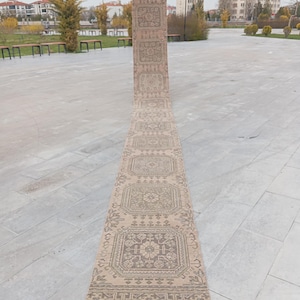 Könnte beinhalten: Ein langer, rechteckiger beigefarbener Teppich mit einem sich wiederholenden geometrischen Muster in Braun- und Grautönen. Der Teppich hat einen roten Fransen am Ende. Der Teppich wird im Freien auf einer Steinoberfläche ausgestellt.