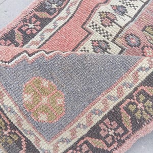 Könnte beinhalten: Ein gefalteter Vintage-Teppich mit floralem Muster. Der Teppich hat eine Farbpalette aus Rosa, Grau, Schwarz und Creme. Das Design umfasst geometrische Formen und florale Motive und schafft eine traditionelle Ästhetik. Die Textur des Teppichs erscheint weich und gewebt.