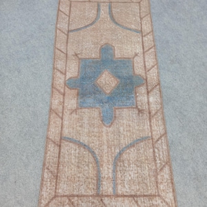 May include: Rectangular rug with a beige and brown border and a central design in blue and beige. The rug features a geometric pattern with a central diamond shape and stylized elements. The rug is placed on a gray surface.