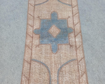May include: Rectangular rug with a beige and brown border and a central design in blue and beige. The rug features a geometric pattern with a central diamond shape and stylized elements. The rug is placed on a grey surface.