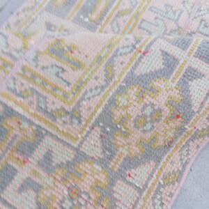 Könnte beinhalten: Aufgerollter, rechteckiger Teppich mit verblichenem, Vintage-Look. Der Teppich zeigt ein sich wiederholendes geometrisches Muster in Rosa, Grau und Gelb. Das Design umfasst florale und rechteckige Formen, die einen traditionellen Look erzeugen.