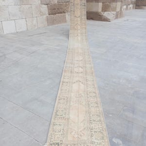 May include: A long, narrow runner rug featuring a faded beige and cream colour palette with intricate, muted grey and light brown geometric patterns along the borders and a central medallion design running the length of the textile piece.
