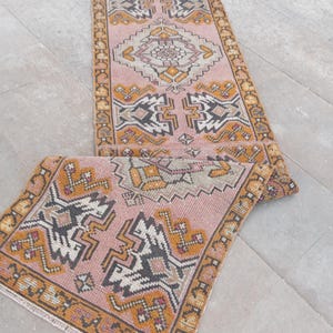 May include: A rectangular runner rug featuring a dusty pink background with geometric patterns in mustard yellow, charcoal gray, and cream. The rug has a decorative border and fringed edges, showcasing a symmetrical design with repeating motifs.