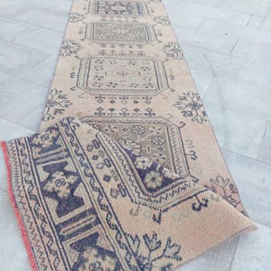 Puede incluir: Una alfombra pasillera larga y rectangular con base beige, decorada con patrones geométricos y medallones centrales en azul oscuro y verde apagado. La alfombra está doblada en un extremo, revelando un borde azul oscuro con motivos florales.