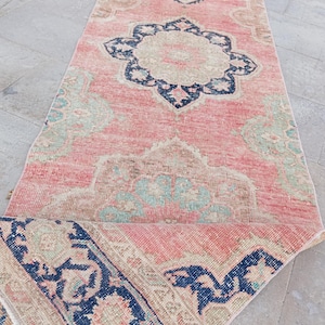 May include: A long, rectangular runner rug featuring a faded coral pink base with intricate floral medallions in navy blue, beige, and soft sage green. The rug displays a distressed texture and a folded corner revealing a dark blue patterned border.