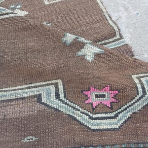 May include: Close-up of a brown, patterned rug with geometric designs. The rug features a central brown field with light blue, pink, and white accents. The design includes star and geometric shapes.