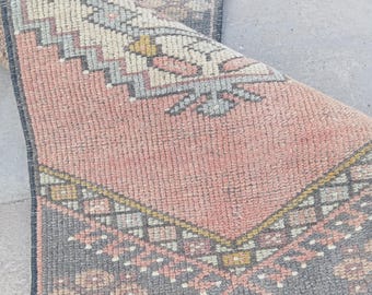 May include: A folded, rectangular rug with a faded, vintage appearance. The rug features a central field of muted coral, bordered by a dark gray pattern with geometric and floral motifs. The design incorporates cream, yellow, and blue accents.
