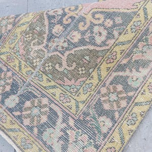 Puede incluir: Una alfombra de estilo vintage con apariencia desgastada. La alfombra presenta un complejo patrón floral en tonos verde oliva, azul marino, rosa pálido y amarillo. Los bordes están deshilachados y la alfombra parece estar doblada sobre sí misma.