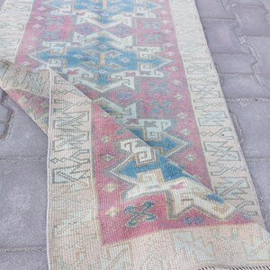 May include: A rectangular runner rug featuring a pink central field with blue and cream geometric patterns. The rug has a cream-colored border with repeating angular motifs and a fringed edge. The corner is folded over to reveal the underside.