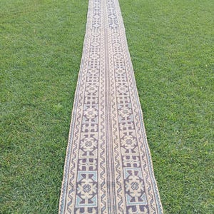May include: A long, narrow rug with a geometric pattern in shades of beige, blue, and brown. The rug is rectangular and appears to be made of woven material. It is laid out on a green grassy surface.
