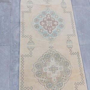 May include: A rectangular, beige-colored rug with an intricate, geometric pattern in shades of blue, green, and brown. The rug features a central medallion design and a border with repeating shapes. The rug is placed on a gray surface.