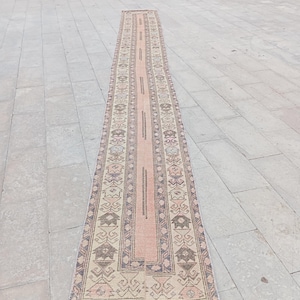 Könnte beinhalten: Ein langer, schmaler Läufer mit einem zentralen blassrosa Paneel, das von komplizierten geometrischen Bordüren in gedeckten Beige-, Braun- und sanften Blautönen flankiert wird. Der Teppich zeigt durchgehend wiederkehrende stilisierte Motive.
