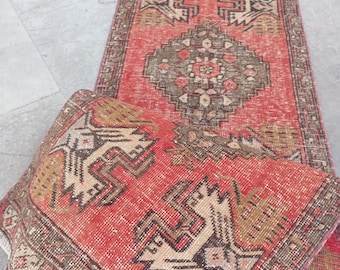 May include: A rectangular runner rug featuring a vibrant red base color with intricate geometric patterns in shades of brown, beige, and cream. The rug displays a central diamond motif and stylized bird-like shapes, finished with a short white fringe.