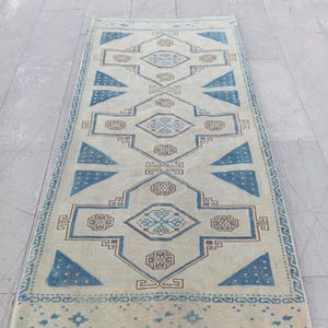 May include: A rectangular runner rug featuring a cream-colored base with a repeating geometric pattern in shades of blue and brown. The design includes central diamond motifs, triangular accents, and intricate border details.