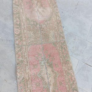 May include: A long, rectangular runner rug featuring a faded, muted colour palette of dusty pink and sage green. The rug displays a repeating geometric medallion pattern with intricate floral motifs throughout the centre and a decorative border.