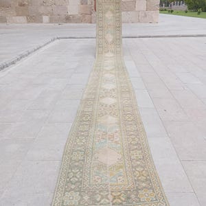 May include: A long, narrow, vintage rug with a repeating geometric pattern in muted tones of beige, green, and blue. The rug is rolled out on a stone surface, showcasing its intricate design and aged appearance. The rug's length is emphasized.