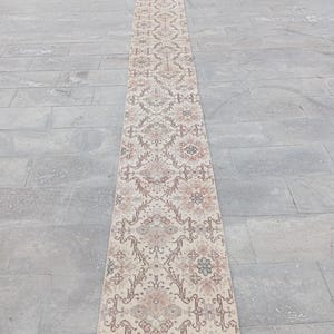 Op de afbeelding: Een lange, rechthoekige loper met een ingewikkeld bloemenpatroon in beige, roze en bruine tinten. Het ontwerp van de loper bevat herhalende bloemmotieven en geometrische vormen. De loper ligt op een grijze tegelvloer.