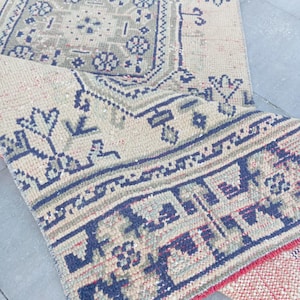 Puede incluir: Una alfombra rectangular con base beige y patrones geométricos intrincados en azul marino y verde salvia apagado. El diseño incluye un medallón octogonal central y motivos florales estilizados. La alfombra está doblada para mostrar el reverso rojo.