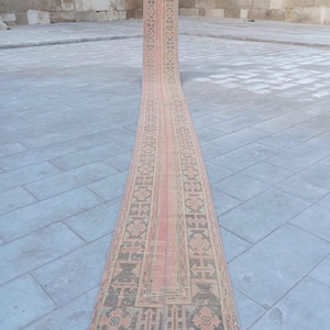 Può includere: Un tappeto da corridoio lungo e stretto con un design persiano tradizionale. Il tappeto è in tonalità di rosa, beige e marrone, con una striscia centrale rosa. Il tappeto ha frange su entrambi i lati.