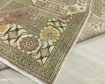 Puede incluir: Una alfombra tejida con una base de color crema y complejos patrones geométricos en tonos rosa empolvado, verde salvia y marrón. La alfombra tiene una esquina doblada que revela un borde con flecos blancos y una cenefa detallada.